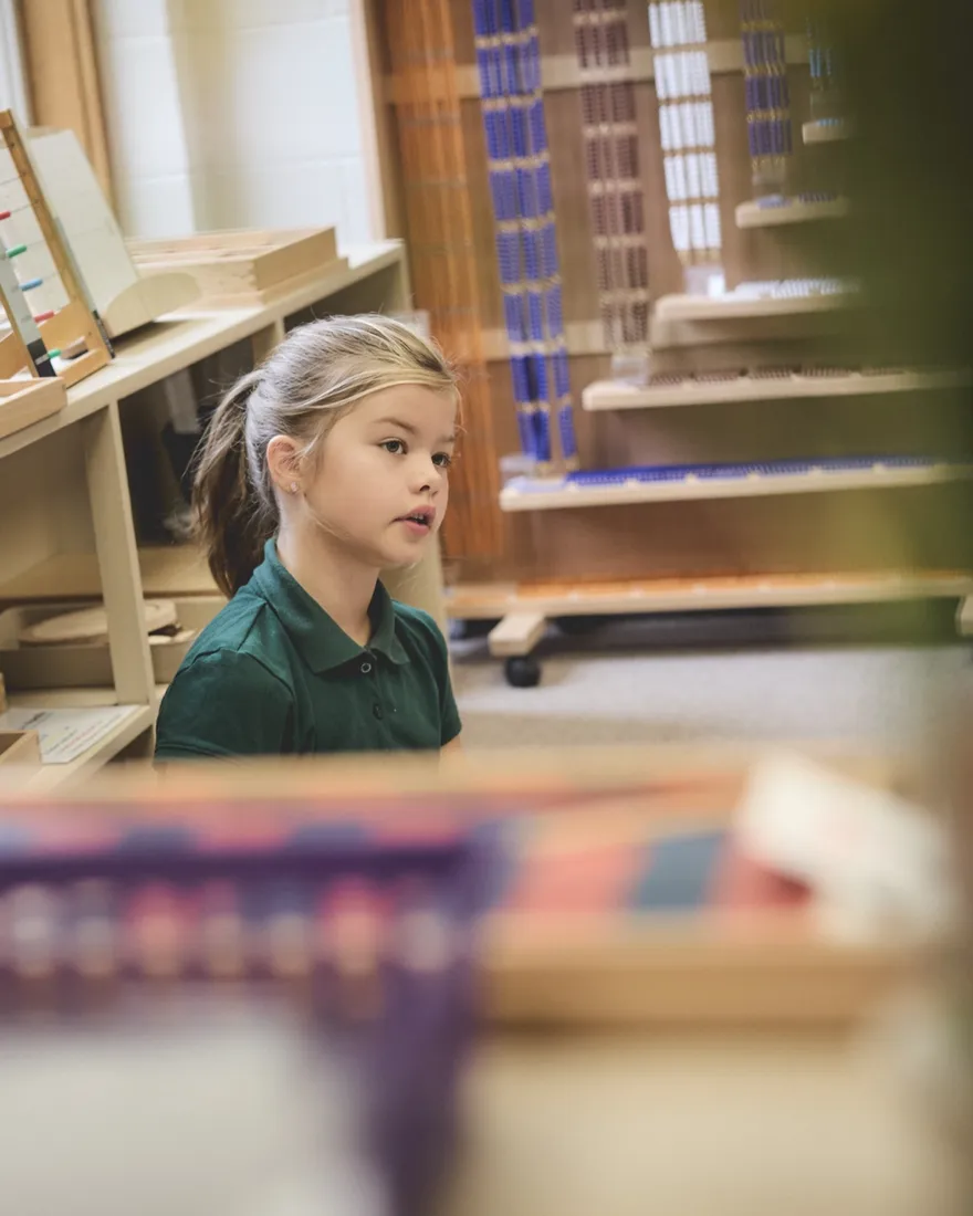 A student working beside shelves of Montessori bead materials in the classroom